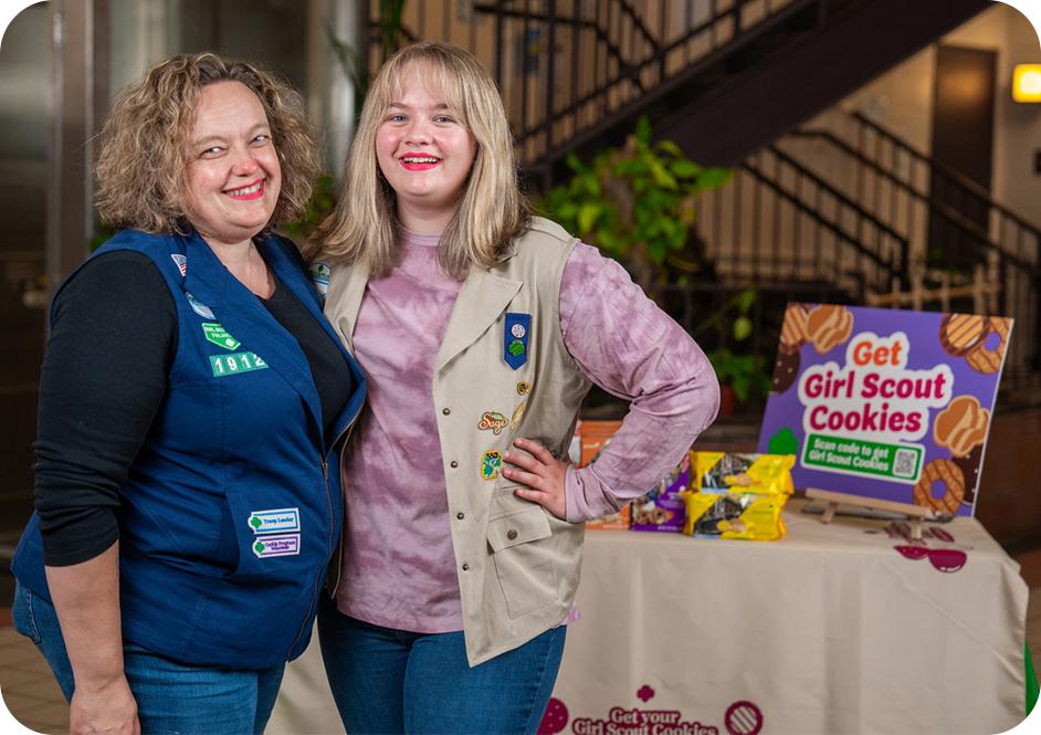 A girl and her troop leader wearing Girl Scouts uniforms standing by table with Girl Scouts cookies
