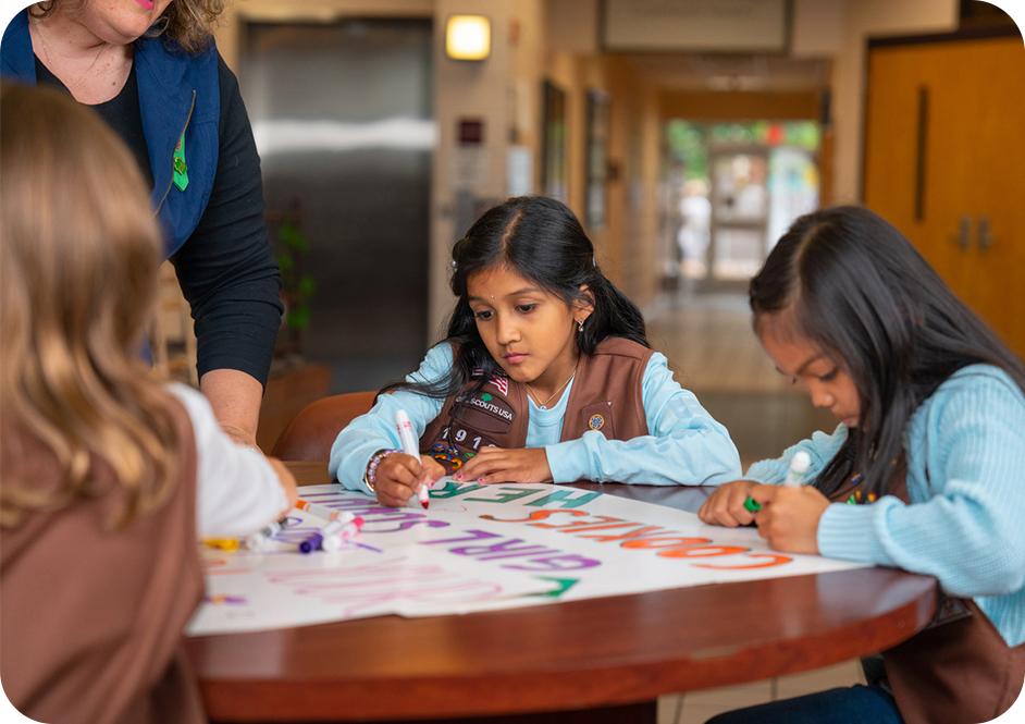 Three girls wearing Brownie Girl Scouts uniforms using markers to make a sign