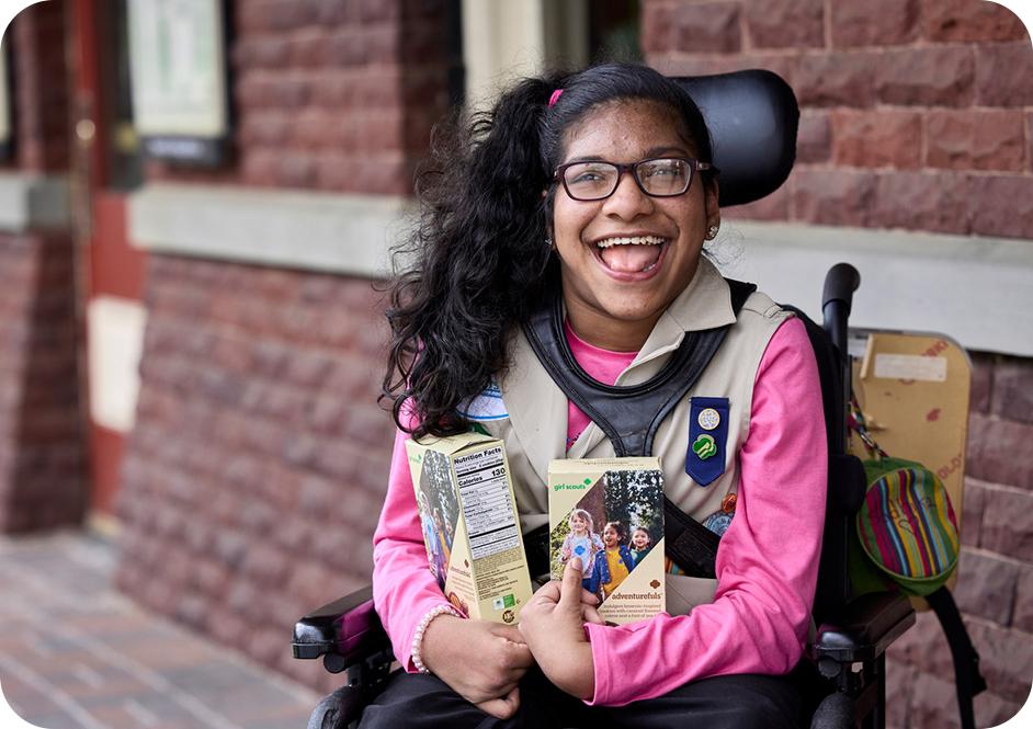 Girl wearing pink long sleeve shirt and tan Girl Scouts uniform holding two boxes of Girl Scouts cookies