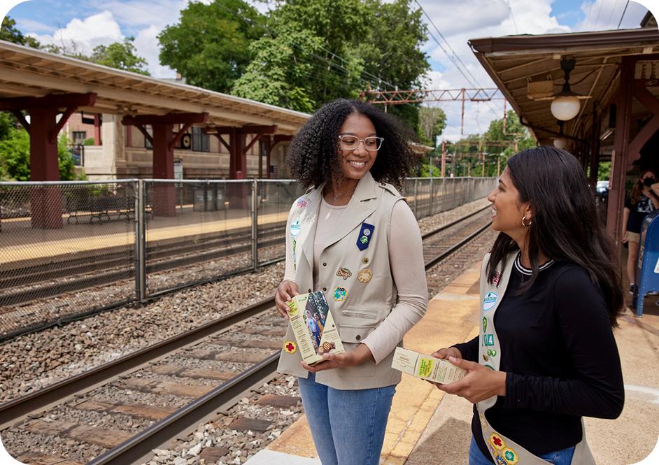 Two girls wearing tan Girl Scouts uniforms and selling Girl Scouts cookies at a train station