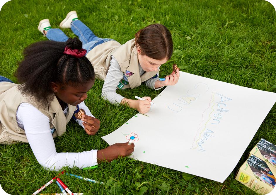 Two girls laying on grass, using markers to create a poster while eating Girl Scouts cookies