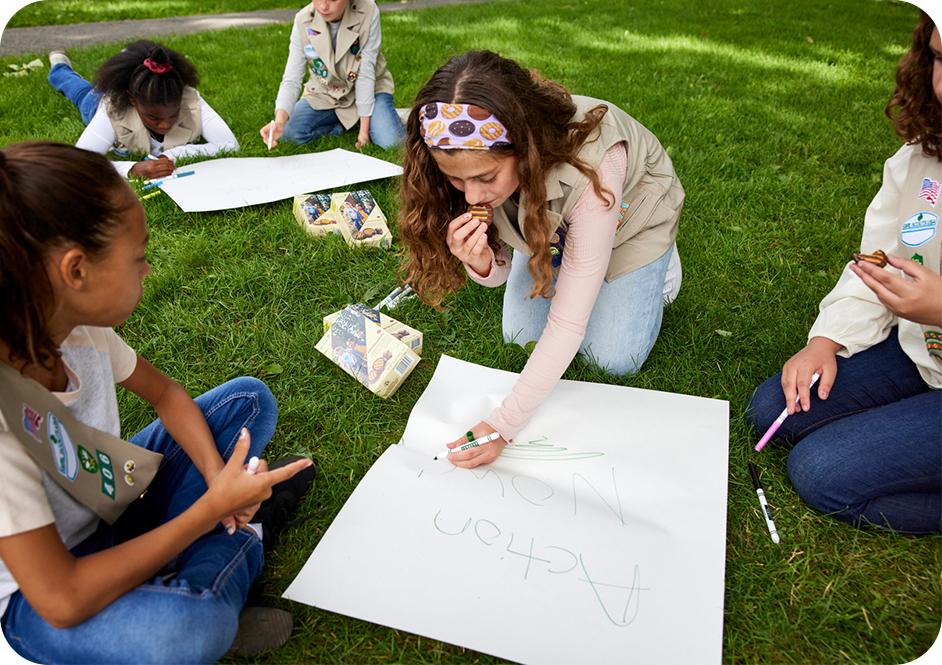 Five girls laying on grass wearing tan Girl Scouts uniforms and using markers to create posters while eating Girl Scouts cookies 