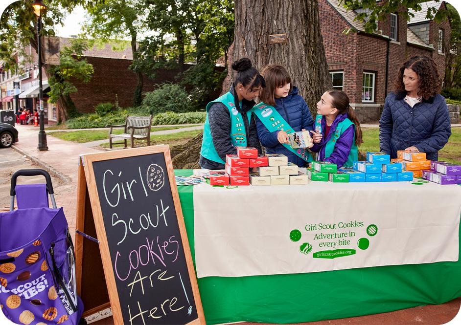 Three girls and a woman wearing green Junior Girl Scouts uniforms selling Girl Scout cookies at a table outdoors