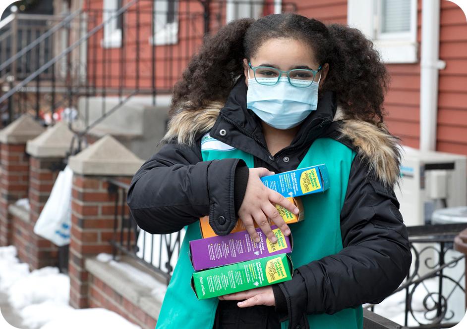 Girl wearing black puffer coat and green Junior Girl Scouts uniform vest and holding 4 boxes of Girl Scouts cookies