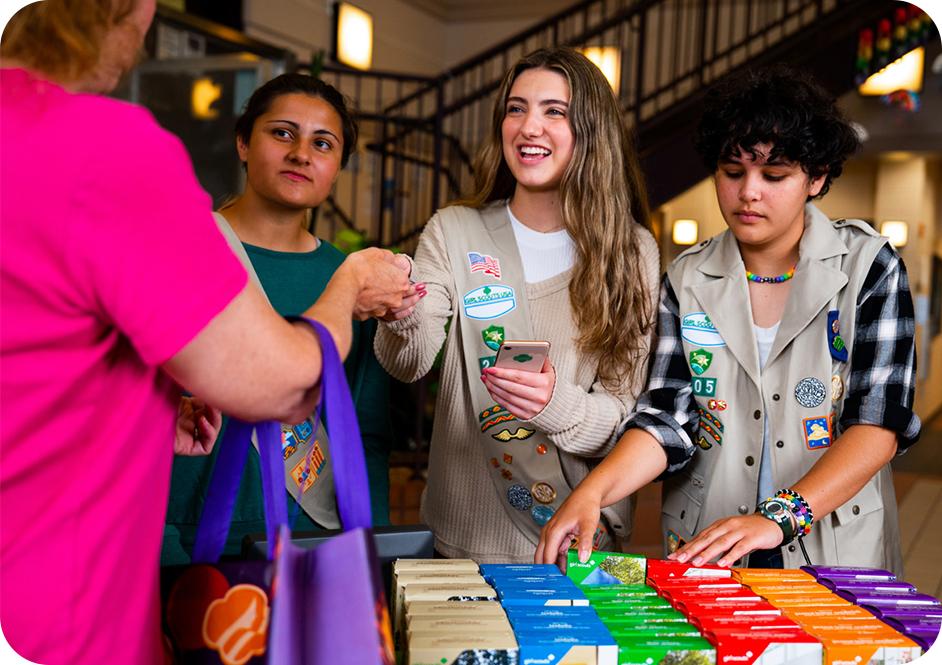 Three girls wearing tan Girl Scouts uniforms selling Girl Scout cookies at a table