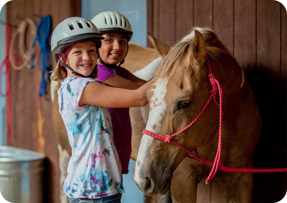 Two girls wearing helmets petting a tan and white horse