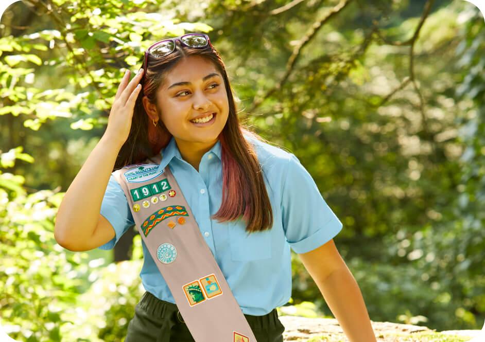 Girl wearing light blue button down short sleeve shirt, green skirt, and tan Girl Scouts uniform sash