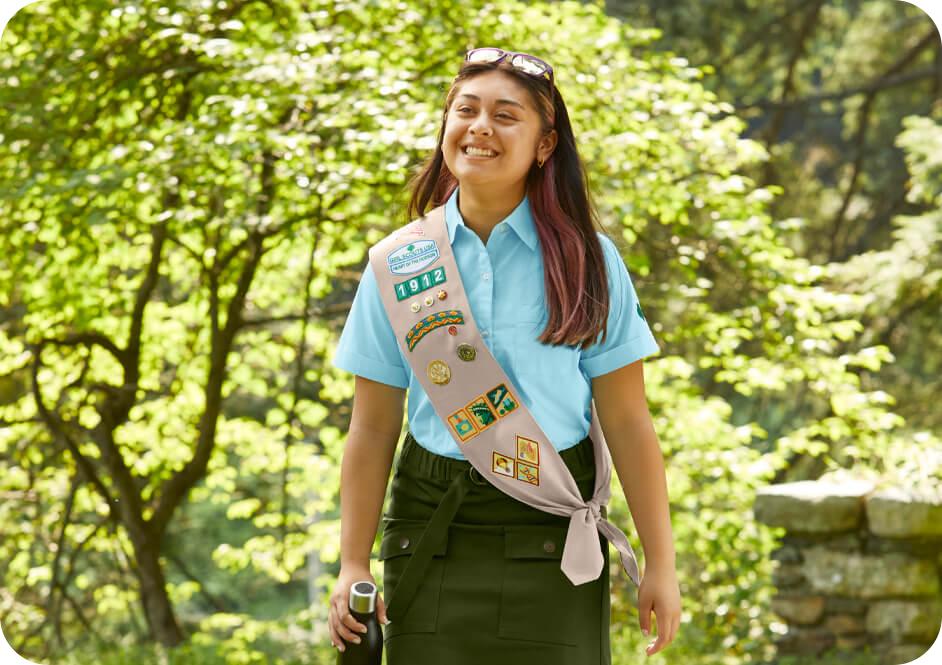 Girl wearing light blue short sleeve button shirt, dark green skirt, and tan Girl Scouts uniform sash