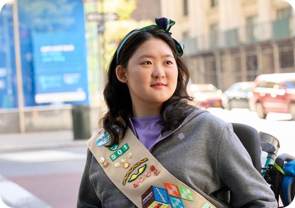 Girl wearing grey Girl Scouts hooded sweatshirt and tan Girl Scouts uniform sash