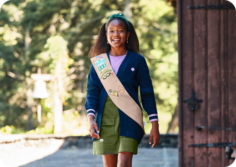 Girl wearing dark blue cardigan sweater, green skirt, and tan Girl Scouts uniform sash