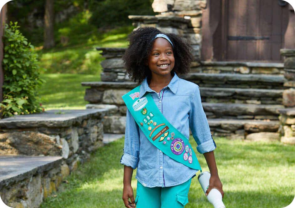 Girl wearing long sleeve light blue chambray shirt with green Girl Scouts uniform sash with many badges on it