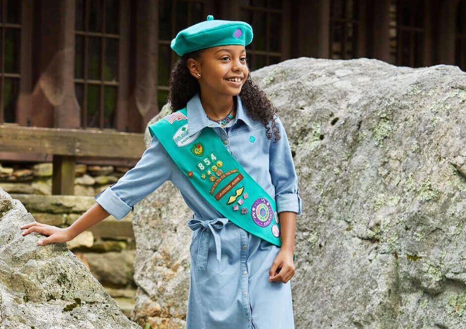 Girl wearing green Girl Scout sash, green beret hat, and blue dress leaning on rock