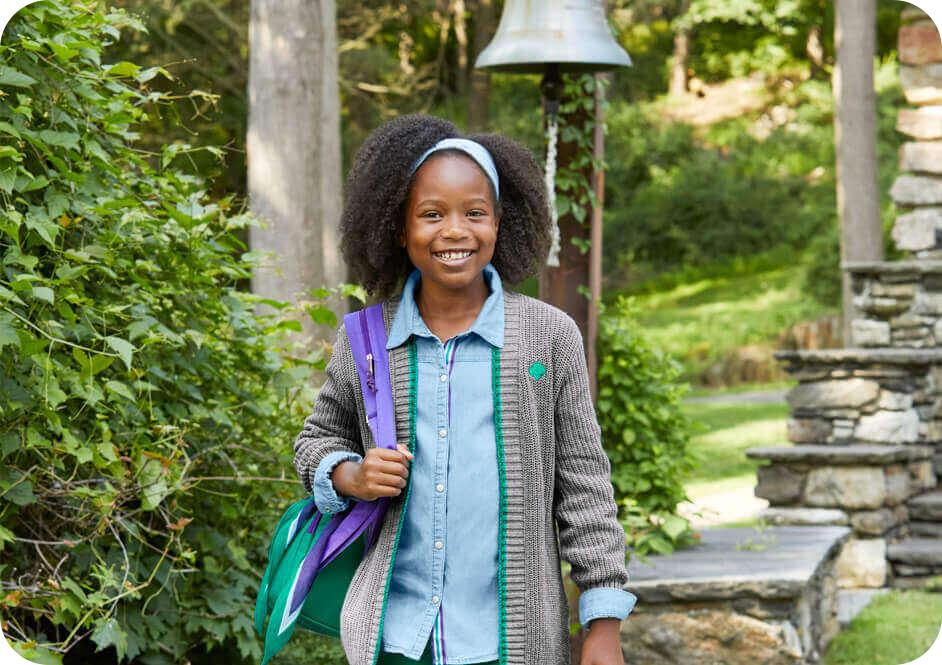 Girl wearing grey Girl Scout cardigan sweater, blue button down shirt, and carrying purple backpack