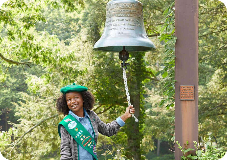 Girl wearing grey Girl Scout cardigan sweater, blue button down shirt, and green beret hat ringing large bell